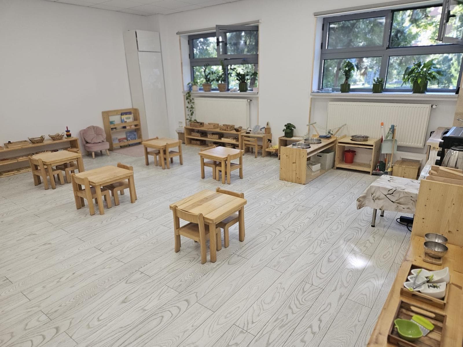 Sală de grupă Montessori goală, mediul pregătit: pardoseală de lemn deschis, mese și scaune la înălțimea copilului, rafturi joase cu materiale ordonate