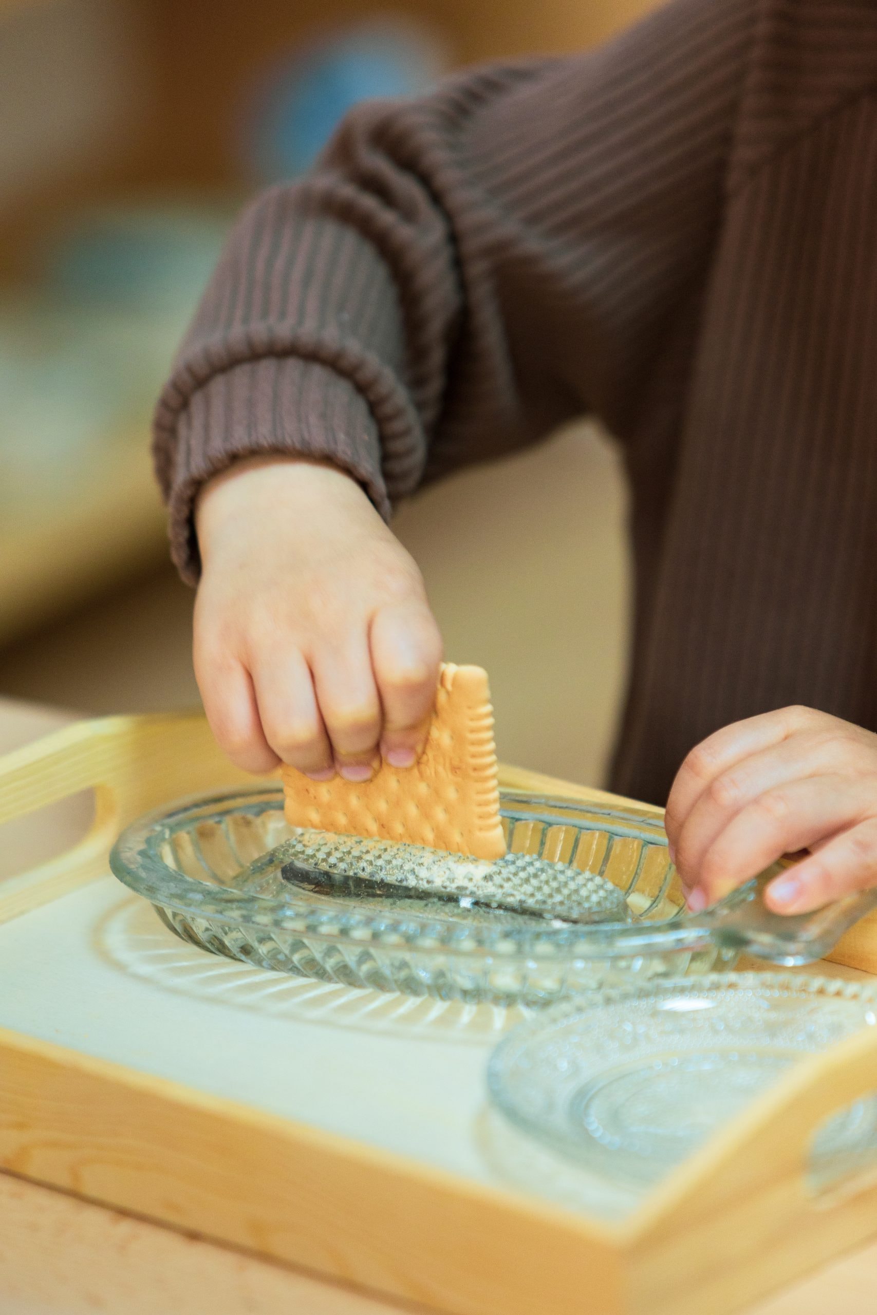 Prim-plan cu mâinile unui copil răzuind un biscuite pe o tavă de sticlă — exercițiu Montessori de viață practică, la Montessori Land