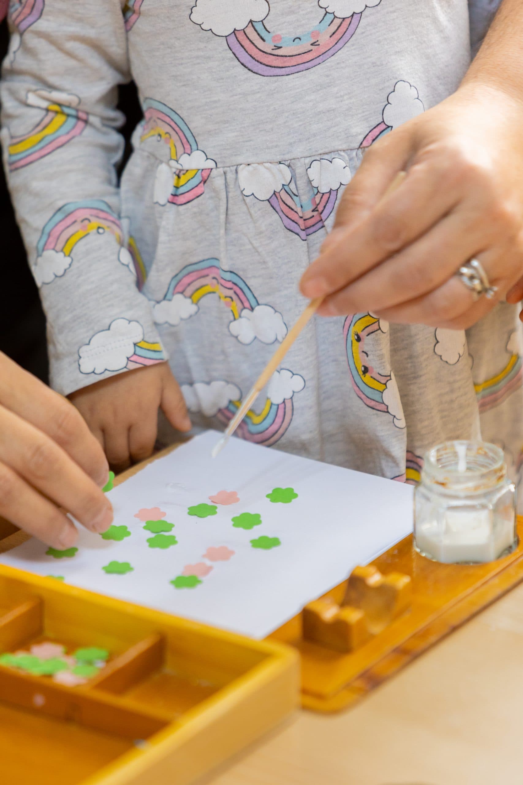 Mâinile unui copil și ale unui adult lipind flori de hârtie pe o tavă de lemn, activitate de artă la creșa Montessori Land