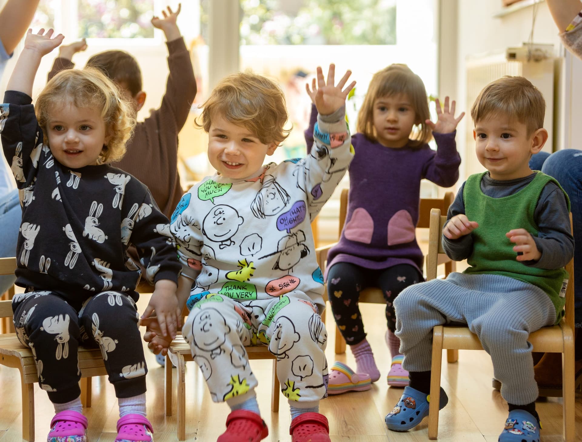 Patru copii mici pe scăunele, cu mâinile ridicate în timpul cercului de dimineață, la creșa Montessori Land