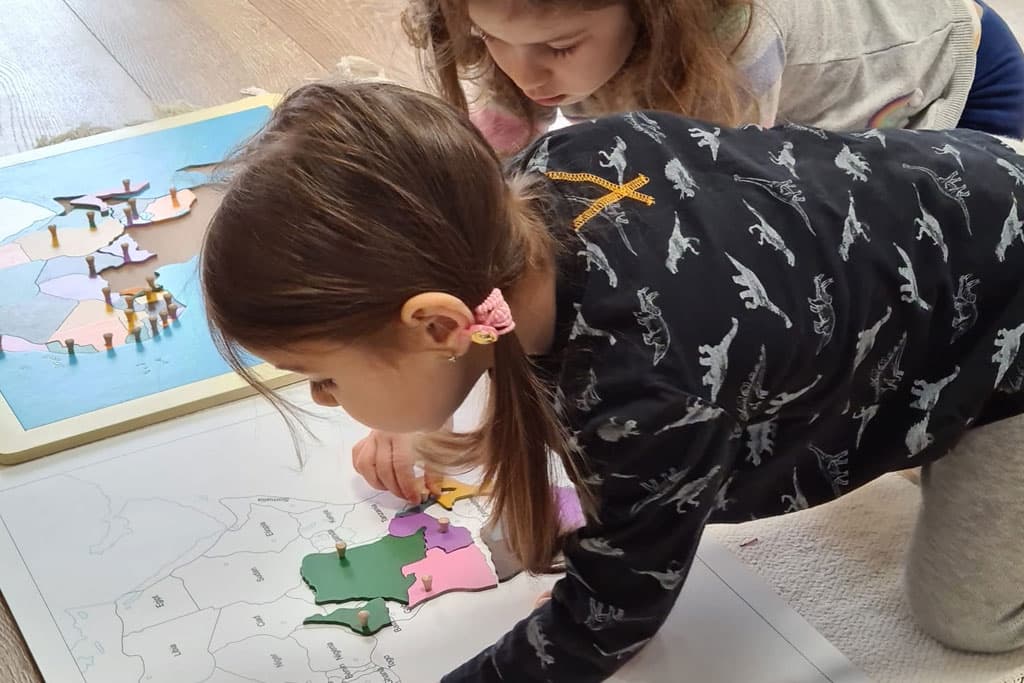 Două fetițe pe podea asamblând o hartă puzzle din lemn cu continentele