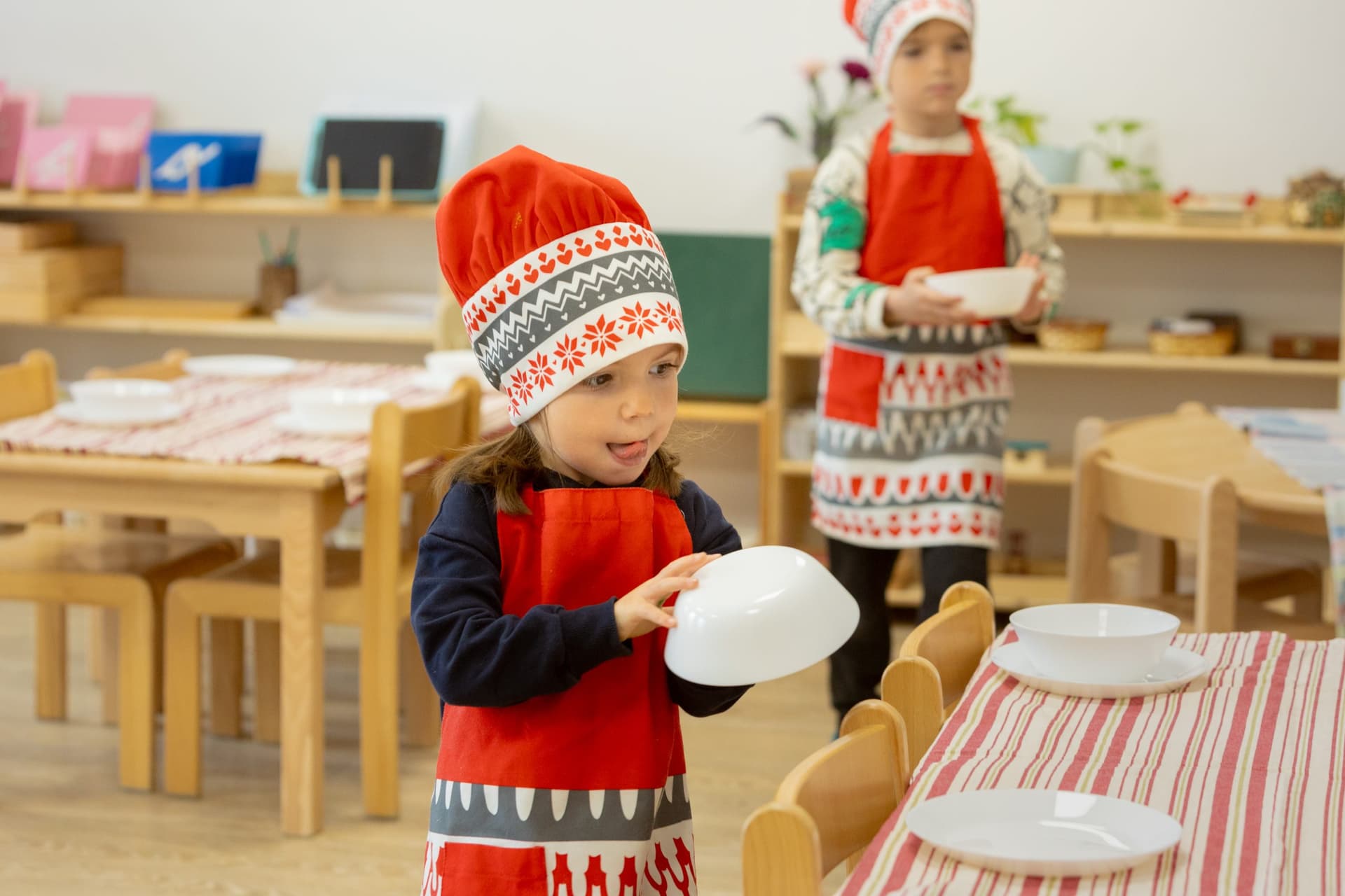 Fetiță cu bonetă de bucătar și șorț roșu ducând un bol către masa de prânz, atelier de gătit la Montessori Land