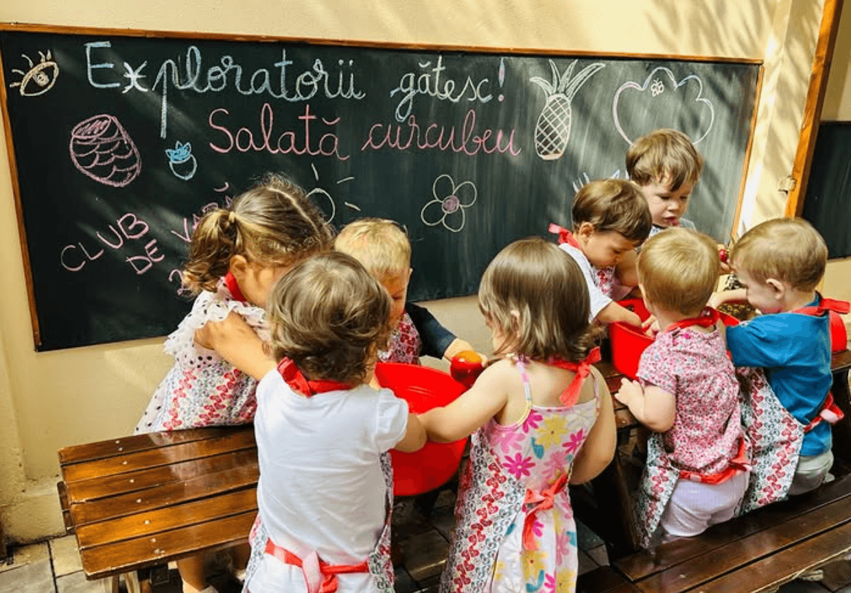 Sală de grupă la sediul Colentina: pardoseală de lemn deschis, mese și scaune la înălțimea copilului, rafturi joase cu materiale Montessori