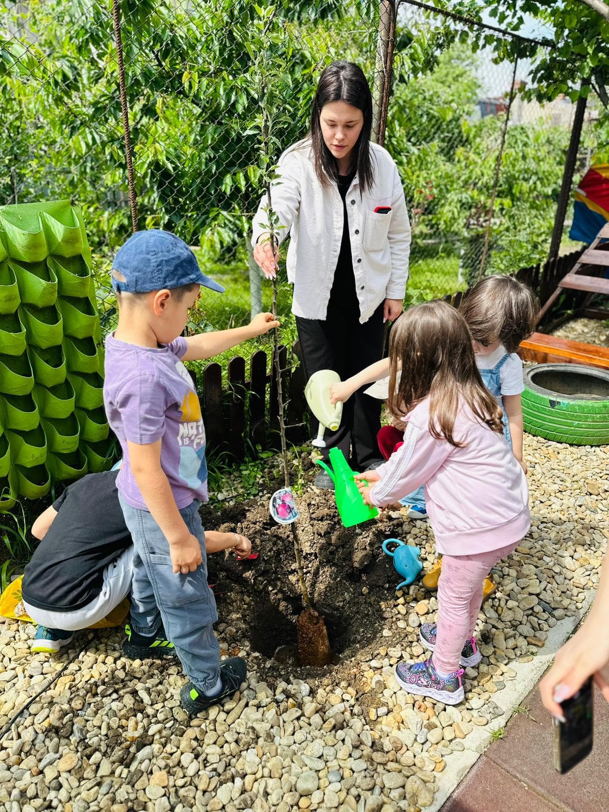 Fetiță în geacă albastră desenând la o masă de picnic în curte, cu domul de cățărat în fundal